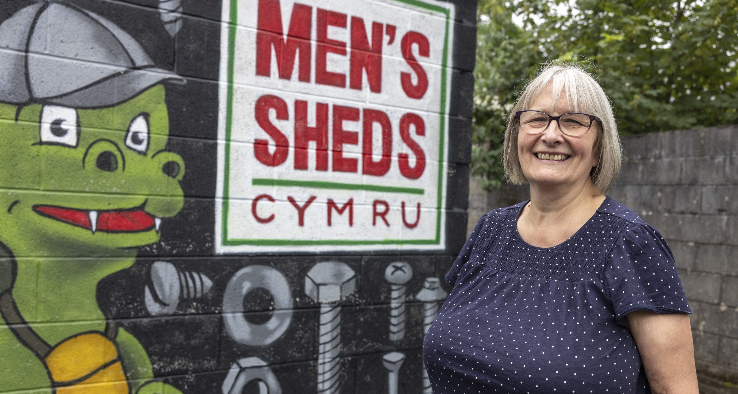 A lady smiling in front of a mural which reads 'Men's Sheds Cymru' next to a dragon wearing a cap.