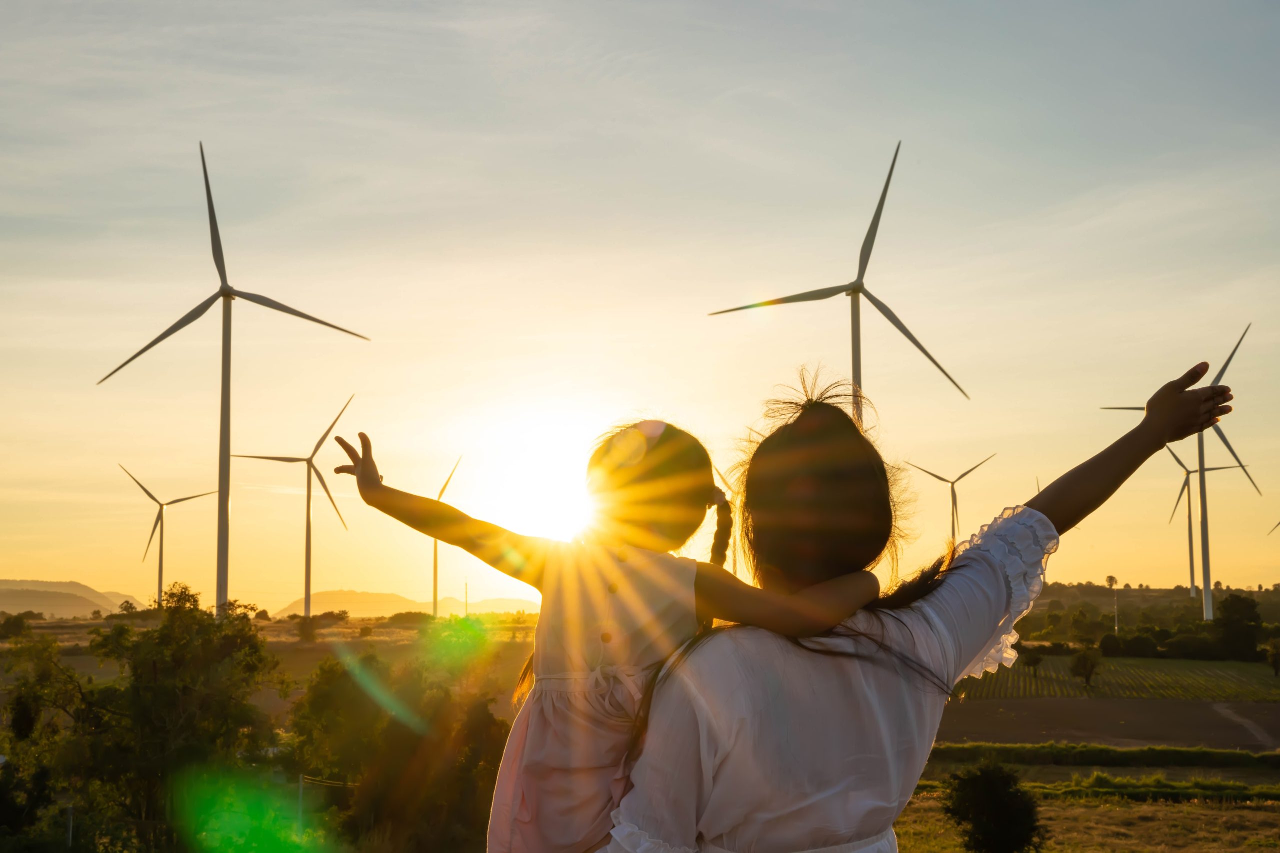 An adult holding a child, with their arms outstretched, both facing wind turbines in the setting sun.